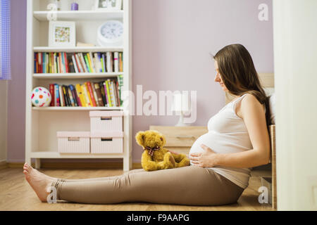 Portrait of beautiful pregnant woman with teddy bear sitting on the floor in bedroom Stock Photo