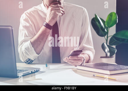 Businessman using mobile phone, working overtime at office desk, multitasking and project deadline concept, retro toned image Stock Photo