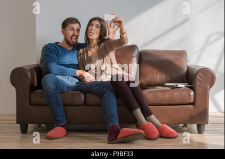Cheerful couple taking a selfie with a smartphone at home sitting on couch Stock Photo