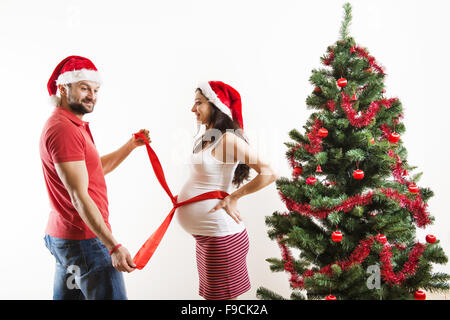 Young couple is standing close to christmas tree. Woman is pregnant and tied with red ribbon. Stock Photo