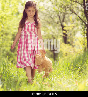 Cute little girl in pink dress is playing with brown teddy in green nature Stock Photo