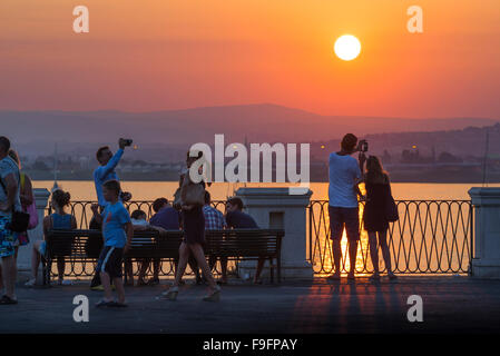 Ortigia Sicily sunset, view of tourists in Ortigia photographing a sunset over the harbour at Syracuse (Siracusa), Sicily. Stock Photo