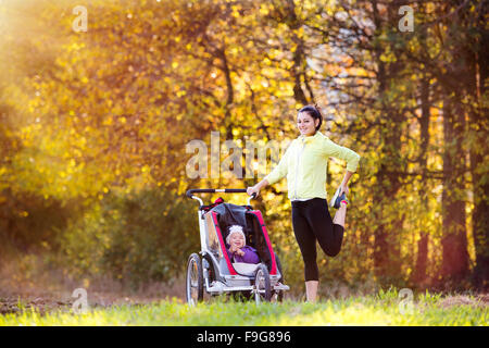 Beautiful young mother with her daughter in jogging stroller running outside in autumn nature Stock Photo