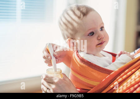 Young mother feeding her little daughter that she has in a baby carrier Stock Photo