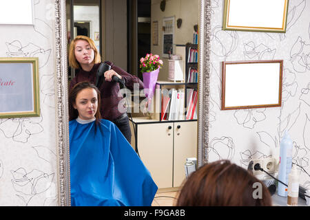 Reflection in Large Mirror of Young Blond Stylist Blow Drying Hair of Brunette Client Wearing Blue Smock in Hair Salon Stock Photo