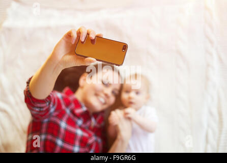 Cute little baby girl and her mother taking selfie on a blanket in a living room. Stock Photo