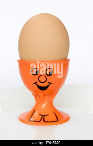 An orange egg cup with a painted smiley face, holding an unbroken brown egg Stock Photo