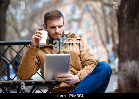 Handsome consentrated bearded young man in coat and jeans drinking coffee and using tablet in open air cafe Stock Photo