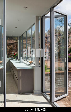 Rear of the house, and bi-fold doors opening from the kitchen onto decking.  Glass panel walls.  A modern family house.