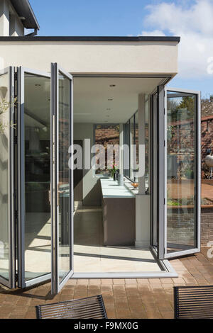 Rear of the house, and bi-fold doors opening from the kitchen onto decking.  Glass panel walls.  A modern family house.