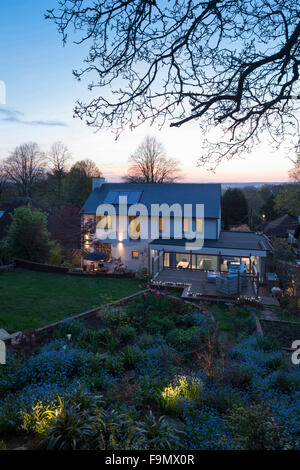House from the rear at dusk showing the terrace, extension and garden. A modern famly house with large full length glass panels. Lights in the twilight. Solar panels on the roof. Stock Photo