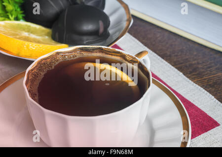 On the table a Cup of black coffee with lemon on the saucer. Next to them are cakes, books. Presents major plan. Stock Photo