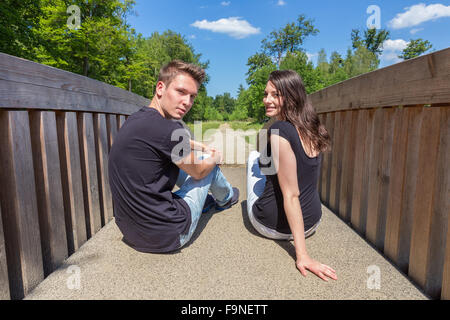 Young caucasian man and woman sitting on wooden bridge looking back Stock Photo