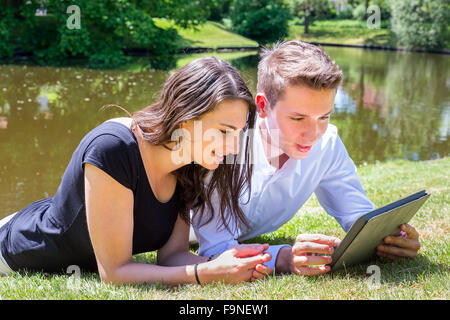 Young couple lying on grass at water reading Ipad Stock Photo