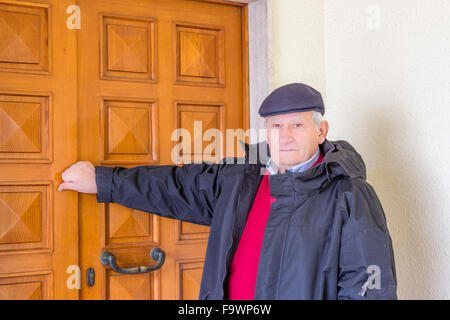 eighty-year-old man in red sweater, dark jacket and cap knocks on the wooden door outside Stock Photo