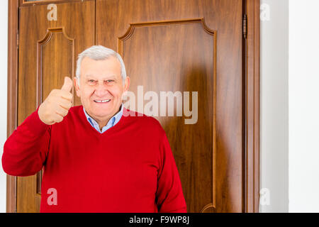 octogenarian happy man in red sweater shows thumb up in front of the door Stock Photo