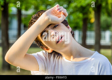 Tired teenager with acne skin holds his head protecting eyes from sunlight with arm on front while showing braces with trees in the background Stock Photo