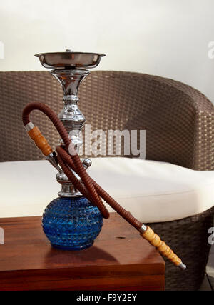 A large, blue, luxury glass hookah sits on a table in the sunshine at an outdoor hookah lounge with a rattan couch. Stock Photo