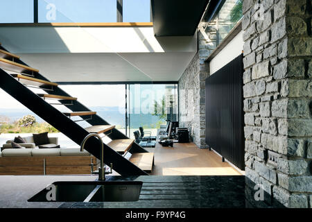 The interior spaces of a modern house on the cliffs overlooking the sea. Large glass panels and walls, and wooden shelves and room dividers.  Floating staircase. Stock Photo