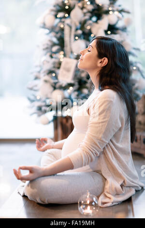 Pregnant woman meditating at home near beautiful decorated Christmas tree, sitting in lotus pose with closed eyes Stock Photo