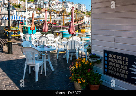 White tables and chairs outside a seafront café offer a splendid view of boats in the picturesque harbour and town of Brixham Stock Photo