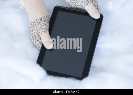 Woman hands in light teal knitted mittens are holding modern tablet pc on snow background. Winter concept. Stock Photo