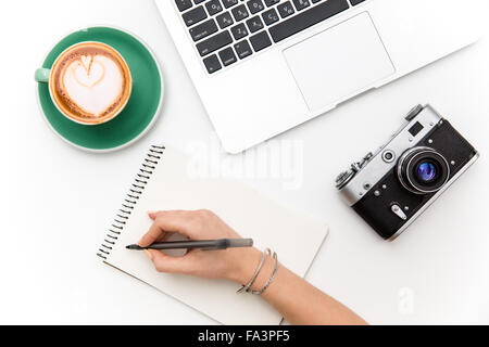 Top view of laptop, old camera, cup of coffee and woman hand writing in notebook over white background Stock Photo