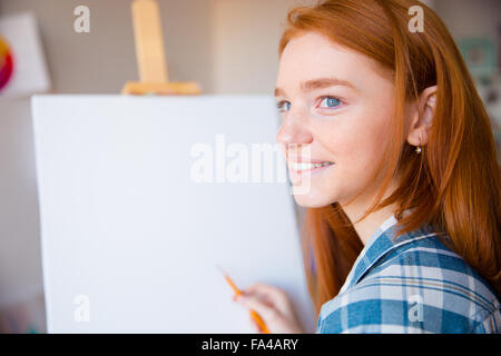 Happy young reahead woman artist in plaid shirt making sketches on canvas in art class Stock Photo