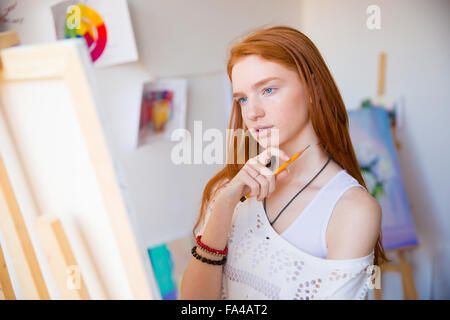 Thoughtful pretty young redhead woman artist thinking and making sketches in art workshop Stock Photo