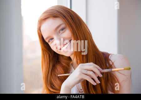 Cheerful happy beautiful redhead young woman with long hair standing near window and holding paintbrush Stock Photo