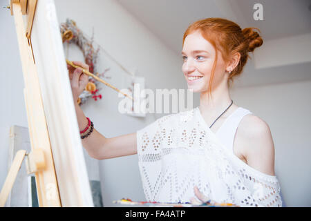 Smiling attractive young woman painter with red hair painting on canvas in artist workshop Stock Photo