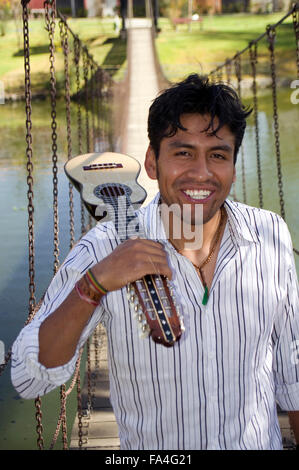 Young man smiling in a striped shirt holds a charango, a traditional Andean string instrument, behind him a bridge over a lake Stock Photo