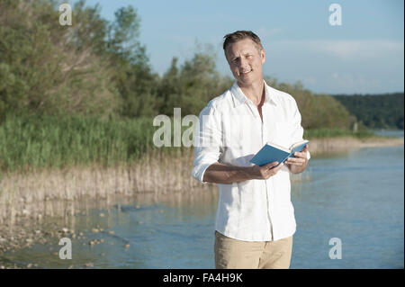 Mature man reading book and smiling at lakeside, Bavaria, Germany Stock Photo