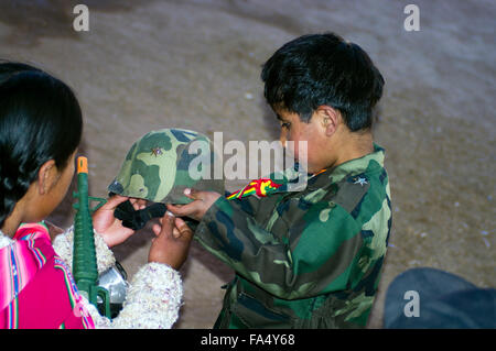 Bolivian boy in soldier's uniform preparing to don his helmet while his mother assists. Stock Photo