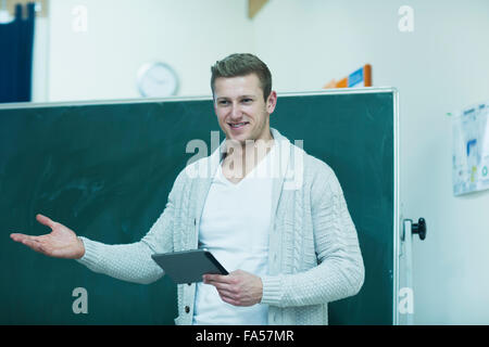 Young male teacher holding a digital tablet and explaining on blackboard in classroom, Baden-Württemberg, Germany Stock Photo