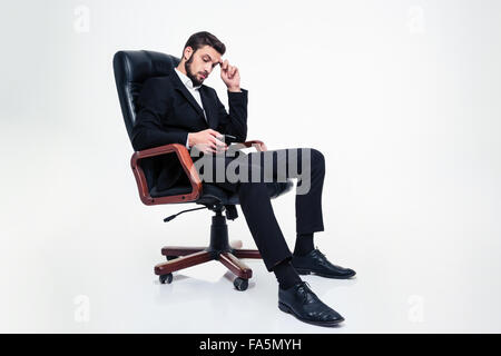 Full length of thoughtful attractive bearded businessman in black suit and shoes sitting in black leather office chair and using Stock Photo