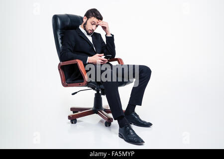 Amazed concentrated businessman with beard in black suit and white shirt sitting in leather office chair and using smartphone ov Stock Photo