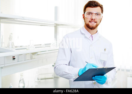 Laboratory worker in protective overall, eyeglasses and gloves making notes Stock Photo
