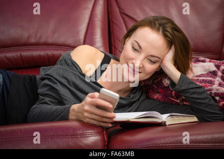 Smiling pretty young redhead woman reading book and using mobile phone at home Stock Photo