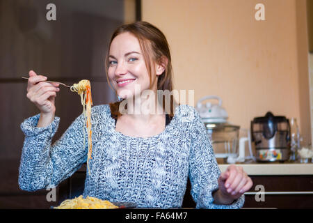 Beautiful cheerful young female in grey knitted sweater  eating pasta in kitchen Stock Photo