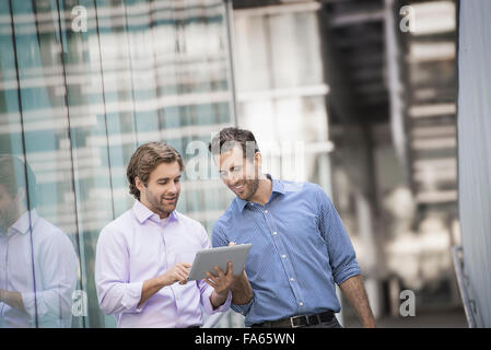 Two men standing outside a large building, one holding a digital tablet. Stock Photo