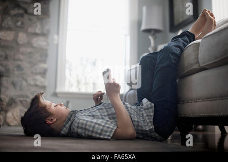 A boy lying on his back on the floor, looking at a digital tablet. Stock Photo