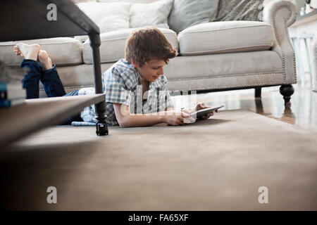 A boy lying on his front on the floor, looking at a digital tablet. Stock Photo