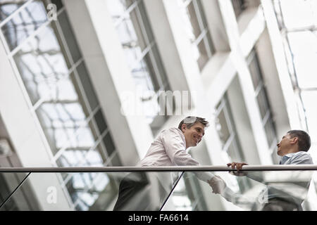 Two men, business colleagues, standing by a railing in an atrium or courtyard, greeting and shaking hands. Stock Photo