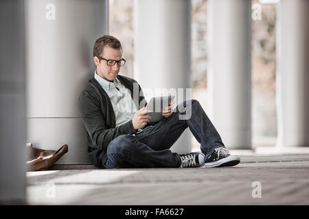 A man sitting leaning against a pillar using a digital tablet. Stock Photo