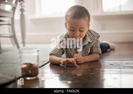 A child lying on his stomach on the floor playing with coins and putting them in a glass jar. Stock Photo