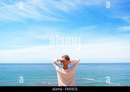 Rear view of a woman standing by sea with hands behind her head, Sesimbra, Lisbon, Portugal Stock Photo