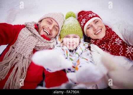 Family of three lying on snow and stretching their hands to the camera Stock Photo