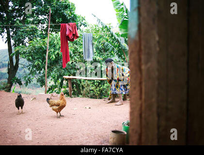 Ugandan Wife sitting on wooden bench in rural yard with chickens and animals Stock Photo