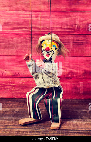 an old marionette with its face painted like a clown being manipulated on a red rustic wooden surface Stock Photo